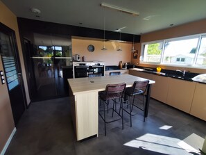 A modern kitchen with a central island, bar stools, a built-in oven, and a large window providing natural light. - 4.5 km from the beach. Beautiful, quiet house. (Saint-Lormel)