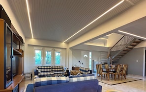 A spacious living area with a sofa set, a television, a dining table with chairs, and a staircase.