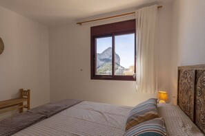A bedroom with a bed, a window with a view of a mountain, a wooden headboard, and a bedside lamp. - Topacio 18 - Costa CarpeDiem (Calpe)