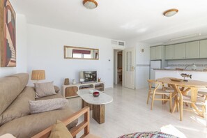 A living room with a sofa, coffee table, and TV, a dining area with wooden chairs and a table, and a kitchen with cabinets and a window. - Topacio 18 - Costa CarpeDiem (Calpe)