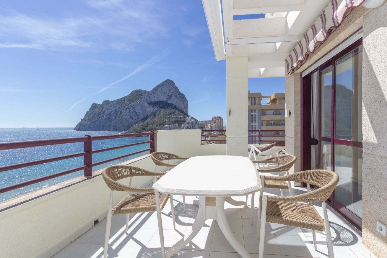 Un balcón con una mesa blanca y sillas de mimbre, con vistas a la costa, una montaña y edificios.