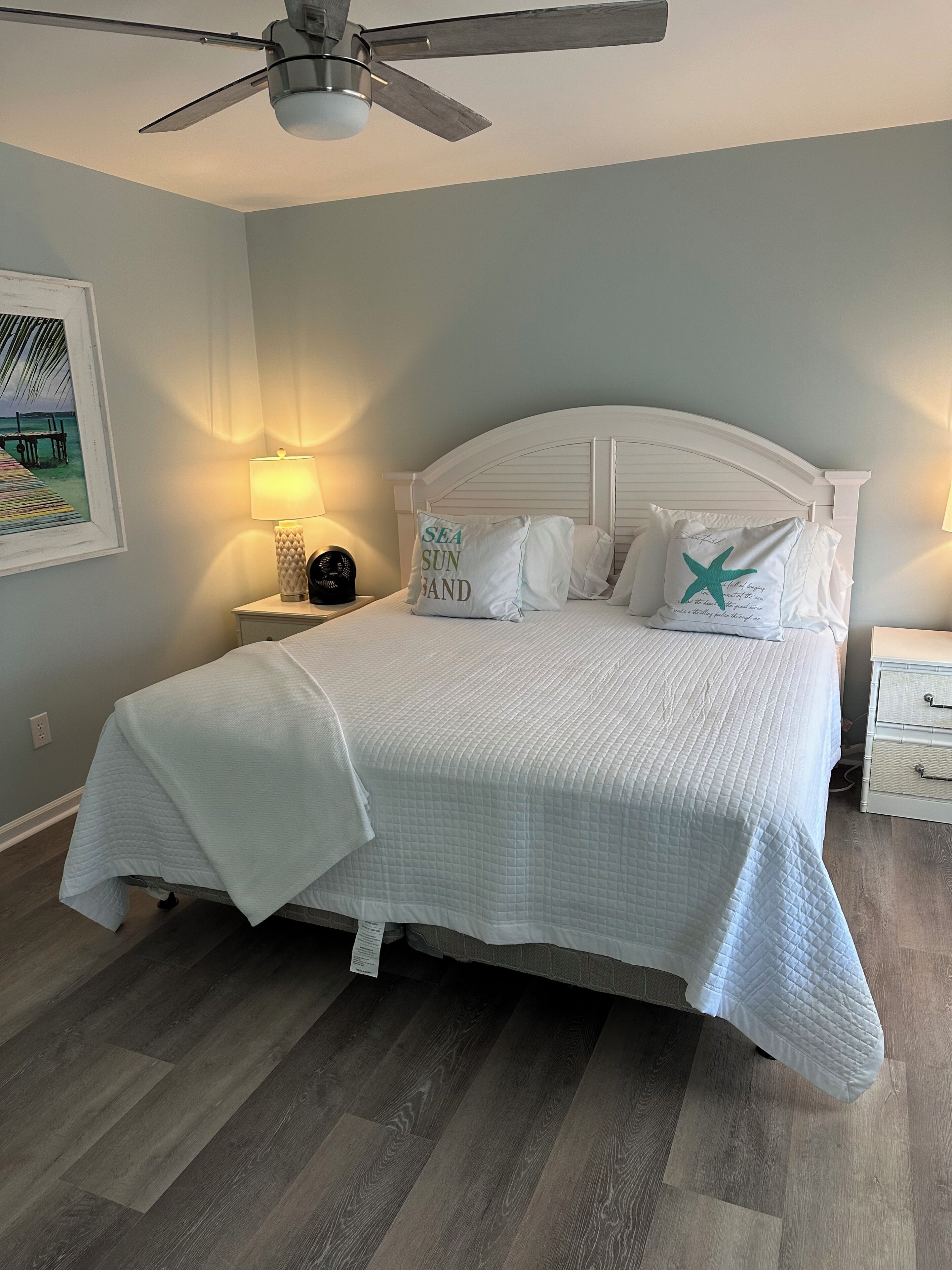 Una camera da letto con un letto, una ventola a soffitto, un comodino, una lampada e un quadro appeso al muro.
