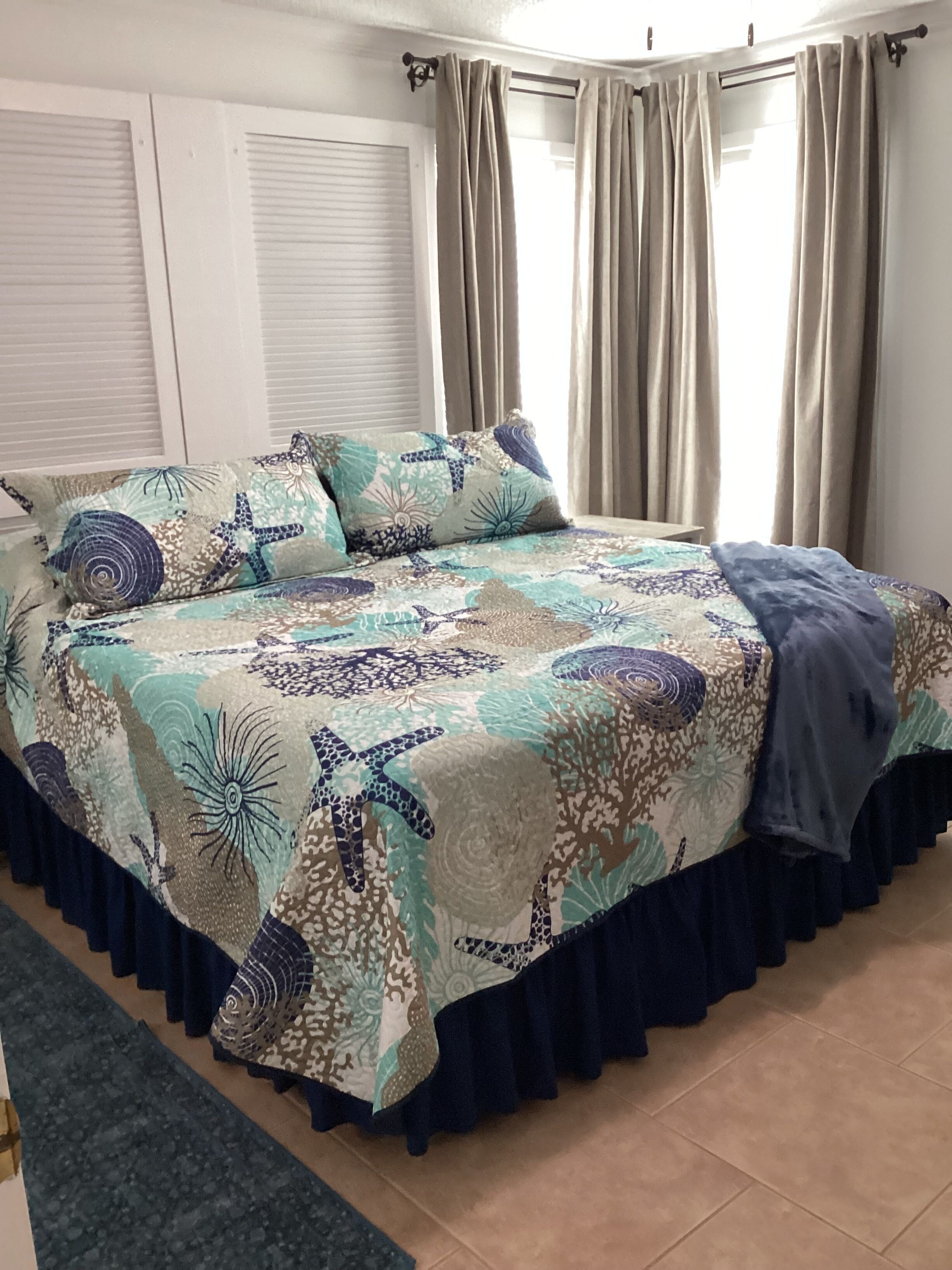 Een bed met een bedsprei met patroon, een donkerblauwe bedrok en een raam met gordijnen.