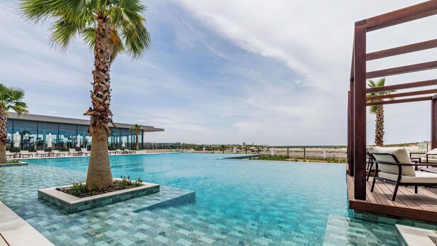 Una piscina con agua cristalina, rodeada de palmeras y sillones de descanso, con vista al horizonte de la ciudad.