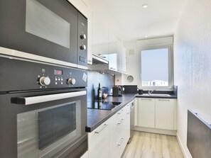A modern kitchen with a built-in oven, microwave, and a window with a view. - Face à l'océan, Appartement avec Garage privatif (Les Sables-d'Olonne)