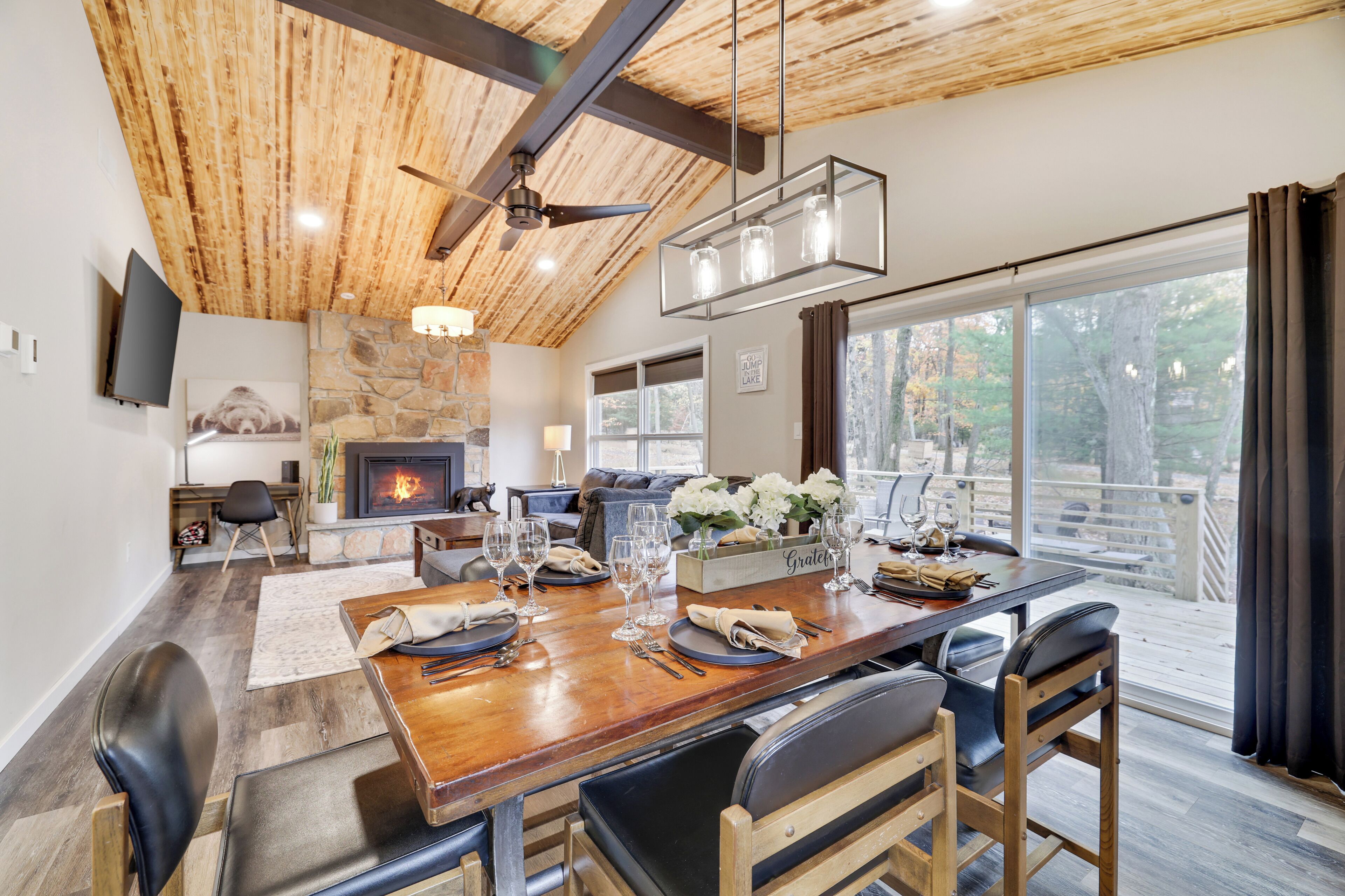 A dining area with a wooden table set for six, a stone fireplace, and a large window with a view of the outdoors.