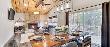 A dining area with a wooden table set for six, a stone fireplace, and a large window with a view of the outdoors.