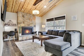 A cozy living room with a stone fireplace, a grey sofa, and a large window with a view of trees. - Harmony Hideaway- Your Perfect Pocono Retreat-Pet Friendly with Hot Tub (Lake Harmony)