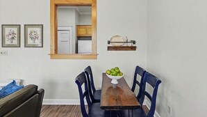 A dining area with a wooden table, blue chairs, a bowl of fruit, a wooden shelf with a plate, and a window with a view into the kitchen. - Full apartment rental with a balcony downtown. Park and walk to everything. (Ocean Springs)