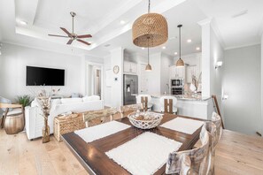 Ein modernes Wohnzimmer mit Essbereich, weißen Wänden, Deckenventilator, Flachbildfernseher und einer Küche im Hintergrund.