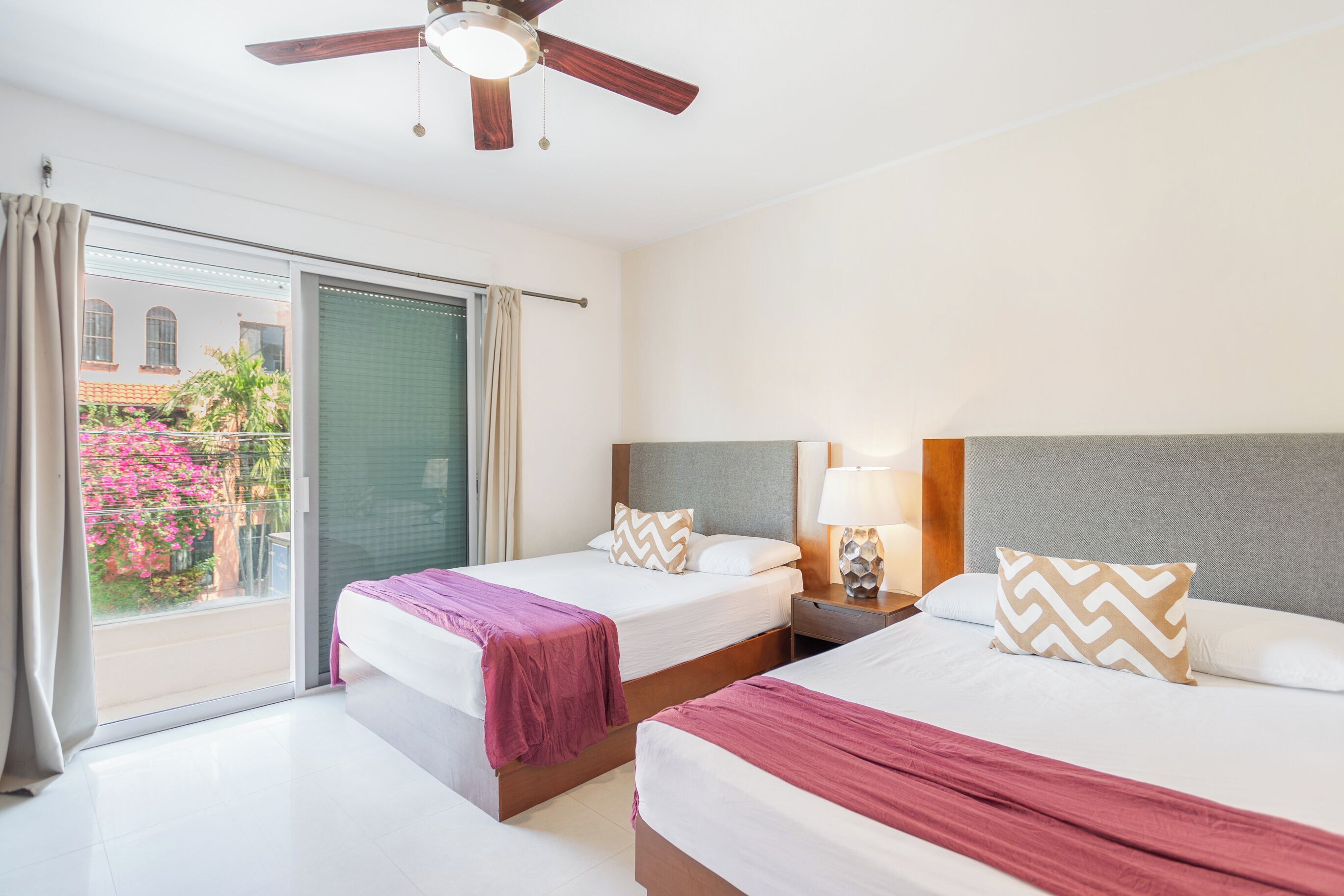 A hotel room with two beds, a ceiling fan, a nightstand with a lamp, and a view of a building and pink flowers through the window.