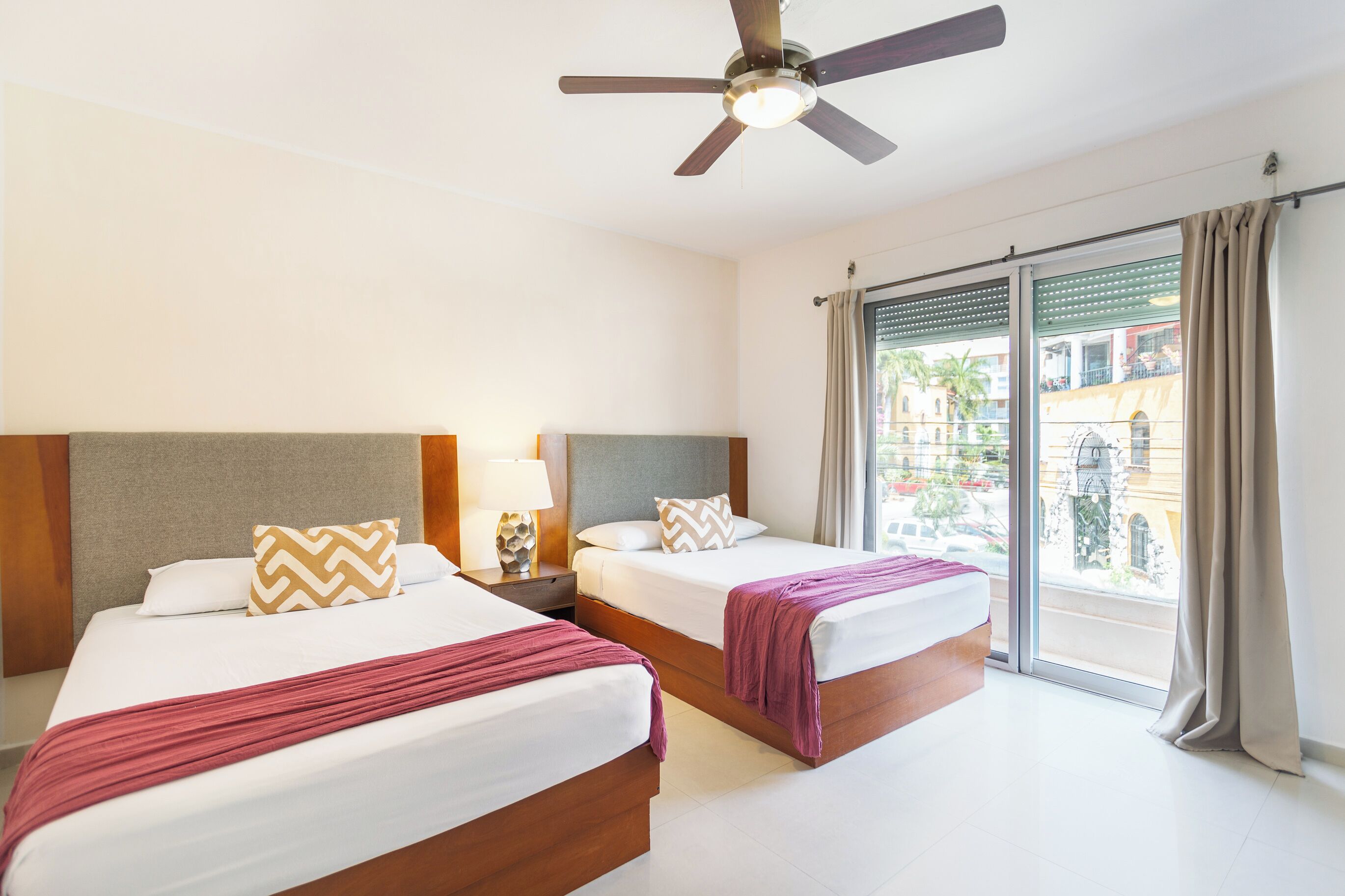 Habitación de hotel con dos camas, un ventilador de techo, una mesita de noche con lámpara y vistas al exterior.
