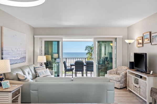 Ein Wohnzimmer mit einer Couch, einem Fernseher, einem Sessel und einem Balkon mit Meerblick.