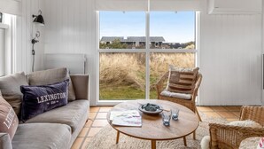 A room with a sofa, a wicker chair, a coffee table, and a large window with a view of buildings and grass. - "Enni" - 350m from the sea by Interhome (Skagen)