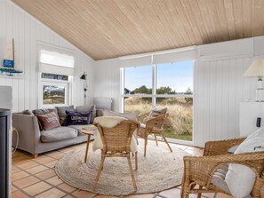 Ein Zimmer mit grauem Sofa, Korbstühlen, einem runden Teppich und einem großen Fenster mit Blick ins Freie.