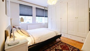 A bedroom with a bed, bedside lamp, window with blinds, wardrobe, and a patterned rug. - Stylish refurbished flat with private garden, near Regent`s Park (Greater London)
