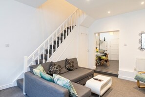 Een woonkamer met een grijze bank, een witte salontafel en een trap naar een andere kamer.