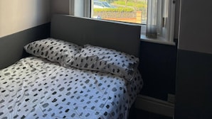 Un dormitorio con cama, ventana con cortinas y vista a edificios y vegetación.