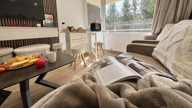 Een moderne slaapkamer met een groot raam, een flatscreen televisie, een ontbijt aan de eettafel en een gezellige leeshoek.