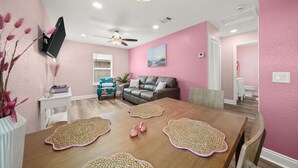 A dining area with a wooden table, woven placemats, a ceiling fan, and a pink wall. - Opalite on Sunset | Pink Beach Retreat (Panama City Beach)