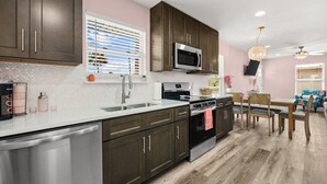 A kitchen with dark cabinets, stainless steel appliances, a white countertop, and a dining area with wooden chairs and a table. - Opalite on Sunset | Pink Beach Retreat (Panama City Beach)
