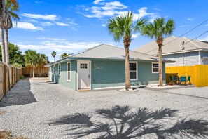 A single-story building with a light green exterior, a white door, and a mailbox. There are two palm trees in front, a gravel driveway, and a yellow fence. - Opalite on Sunset | Pink Beach Retreat (Panama City Beach)