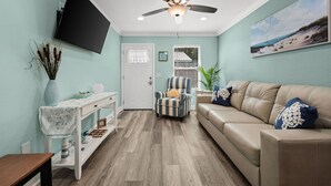 A living room with a ceiling fan, a TV mounted on the wall, a beige sofa, a striped armchair, a small table with a vase, and a window with blinds. - Sun Kissed Cheeks w/ Fire Pit & Beach Gear (Panama City Beach)