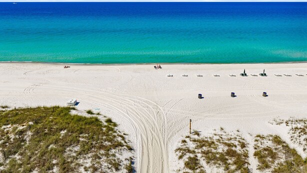 A beach with a clear view of the ocean, a sandy area with some structures, and a grassy foreground. - Sun Kissed Cheeks w/ Fire Pit & Beach Gear (Panama City Beach)
