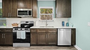 A modern kitchen with dark wooden cabinets, stainless steel appliances, and a window with a view of greenery. - Sun Kissed Cheeks w/ Fire Pit & Beach Gear (Panama City Beach)