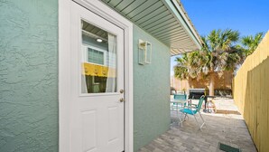 A house with a white door, a small window, a patio with a table and chairs, and a palm tree in the background. - Sun Kissed Cheeks w/ Fire Pit & Beach Gear (Panama City Beach)
