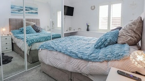 Een slaapkamer met een bed, nachtkastjes, een spiegel, een plafondventilator, een televisie en een raam met zonwering.