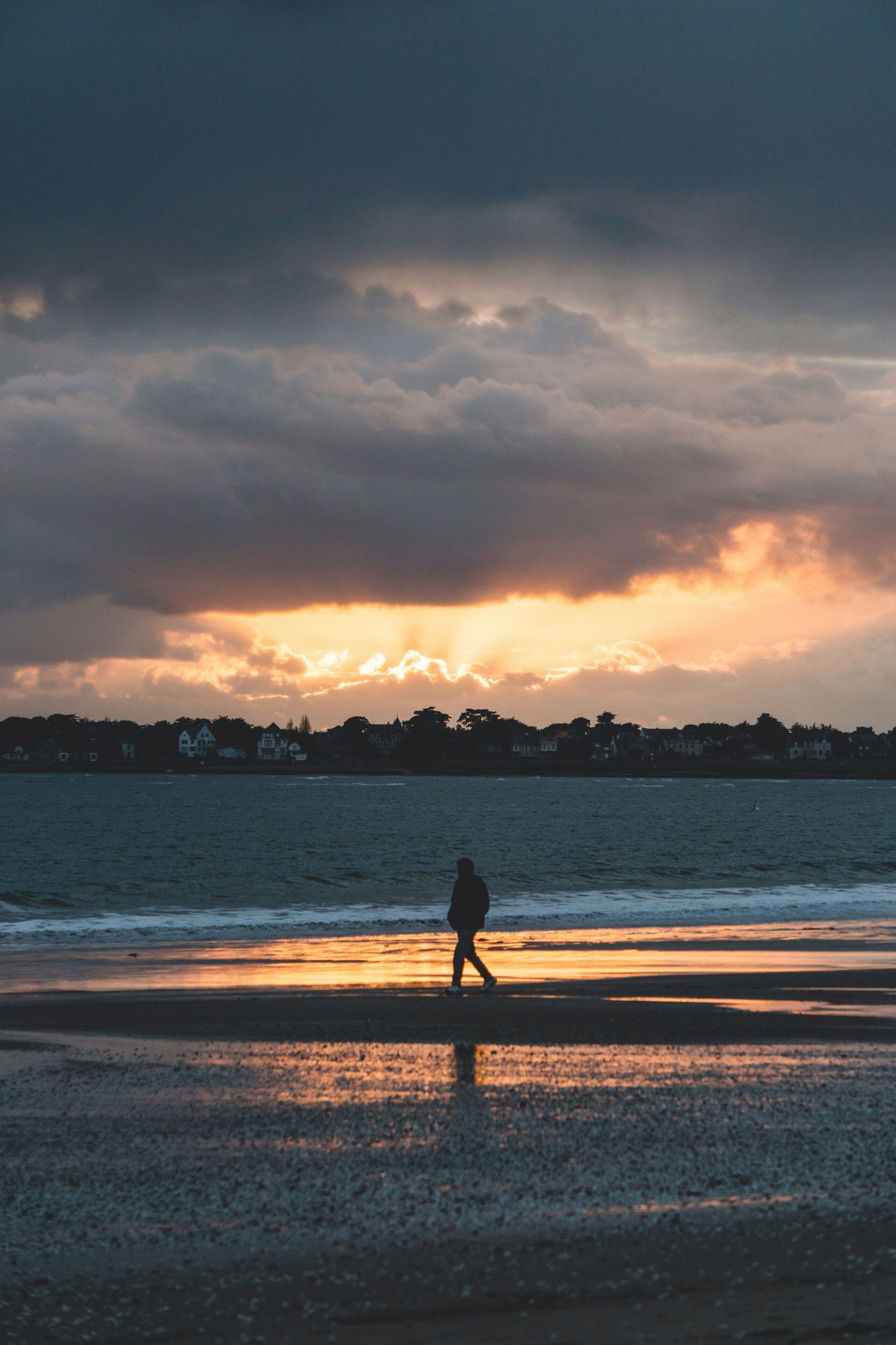 Silueta de una persona caminando en la playa al atardecer, con cielo nublado y casas al fondo.