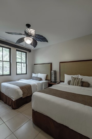 Ein Hotelzimmer mit zwei Betten, einem Deckenventilator, einem Nachttisch, einer Lampe und großen Fenstern.