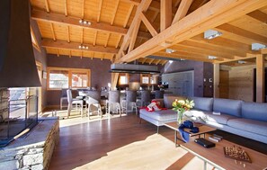 A spacious living area with a high wooden ceiling, a fireplace, and a modern kitchen in the background. - Chalet 270 m2 | Hammam, gym, balcon + vue montagne (Les Deux Alpes)