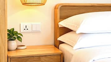 Cama bem arrumada com lençóis brancos, mesa de cabeceira de madeira com um vaso de planta e luminária fixada na parede.