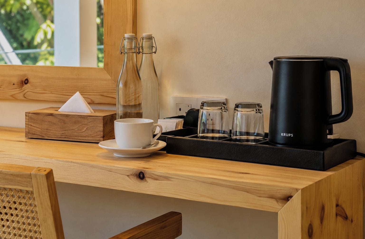 A wooden table with a white cup, a black coffee maker, and two glass bottles.