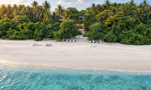 A beach with clear turquoise water, white sand, and palm trees. There are lounge chairs and a building in the background. - ODI Beachfront (Kamadhoo)