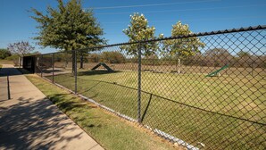 A fenced outdoor area with a grassy field, trees, and a playground. - "Pooling Around " in Aggieland" (College Station)