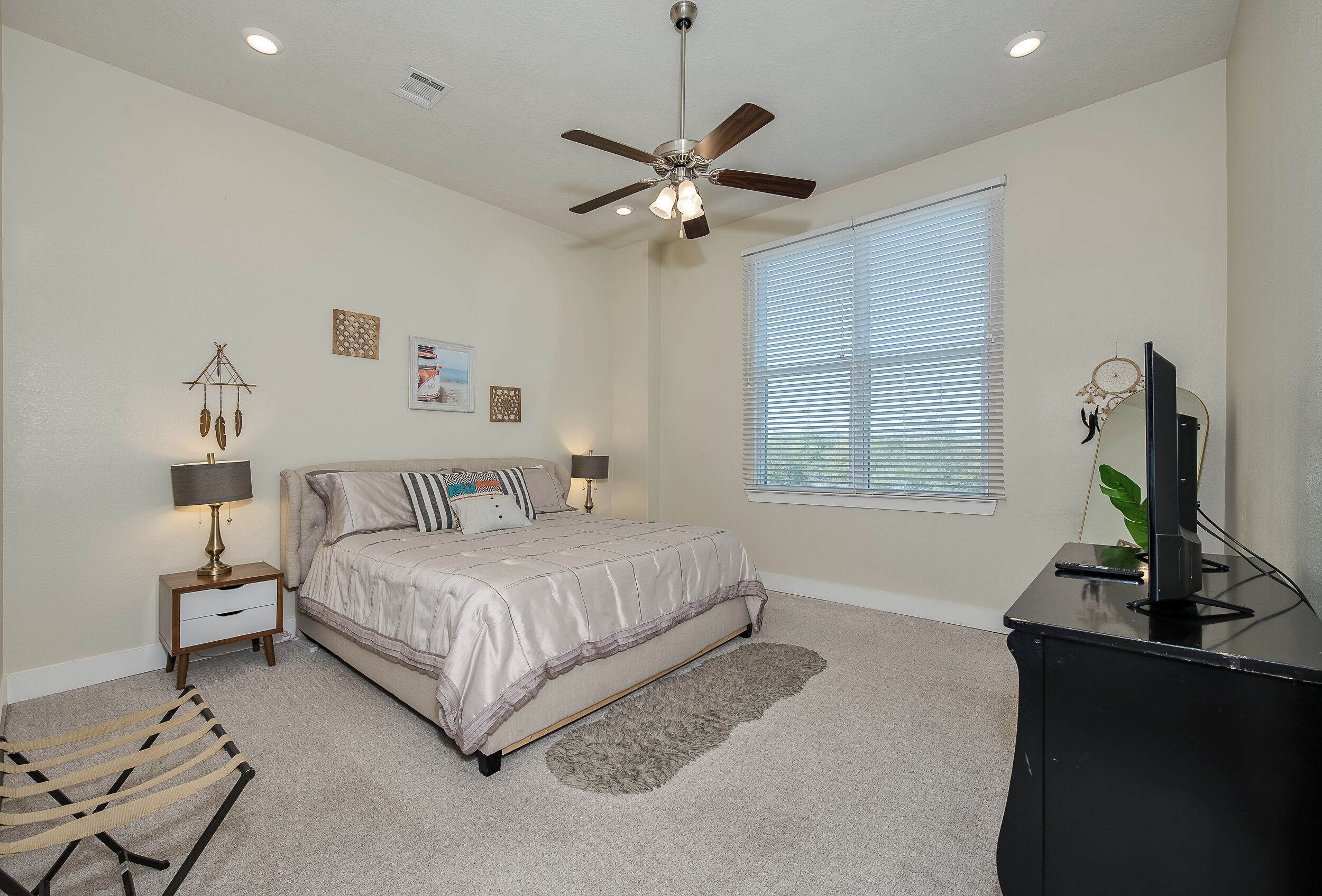 A bedroom with a bed, a ceiling fan, a nightstand with a lamp, a desk with a monitor, and a window with blinds.
