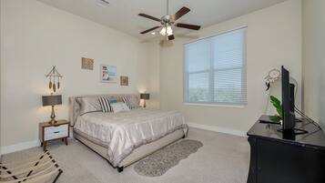 A bedroom with a bed, a ceiling fan, a nightstand with a lamp, a desk with a monitor, and a window with blinds.