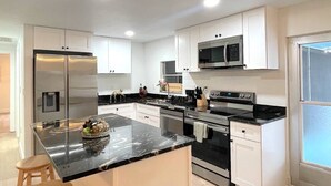 Une cuisine moderne avec des armoires blanches, un réfrigérateur en acier inoxydable, un four à micro-ondes et une cuisinière. On y trouve un tabouret en bois et une poubelle.
