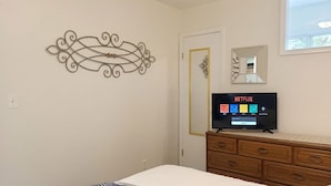 Une chambre à coucher comprenant un lit, une commode en bois, un téléviseur fixé au mur diffusant Netflix, une œuvre d’art décorative en métal accrochée au mur, et une porte dotée d’un cadre doré.