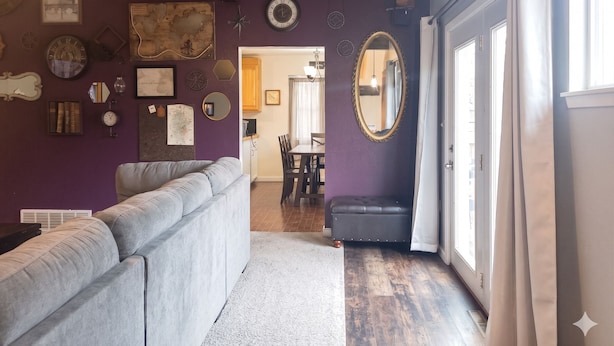 Un salon avec un canapé gris, un coin repas avec une table et des chaises, et un couloir menant à une porte.