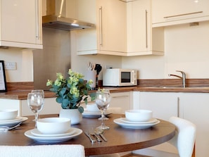 Una cocina moderna con armarios blancos, una campana extractora de acero inoxidable, un microondas y un fregadero. Hay una mesa de comedor con platos, tazones y copas de vino blancos. Una planta en maceta está sobre la mesa.