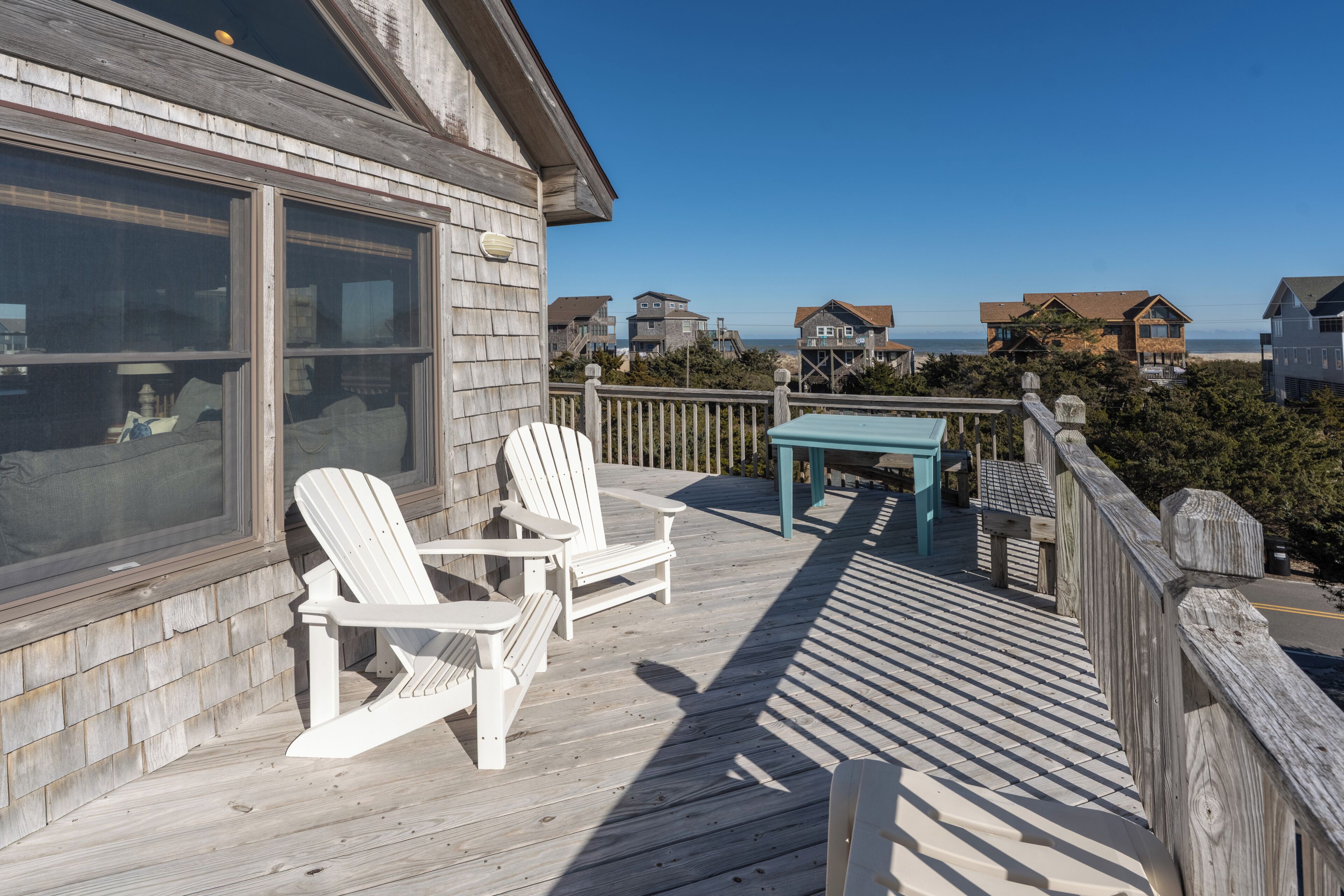 A wooden deck with Adirondack chairs and a table, overlooking a beach with houses in the background.