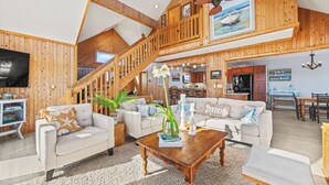 A spacious living area with a high ceiling, wooden beams, a staircase, and a dining area.