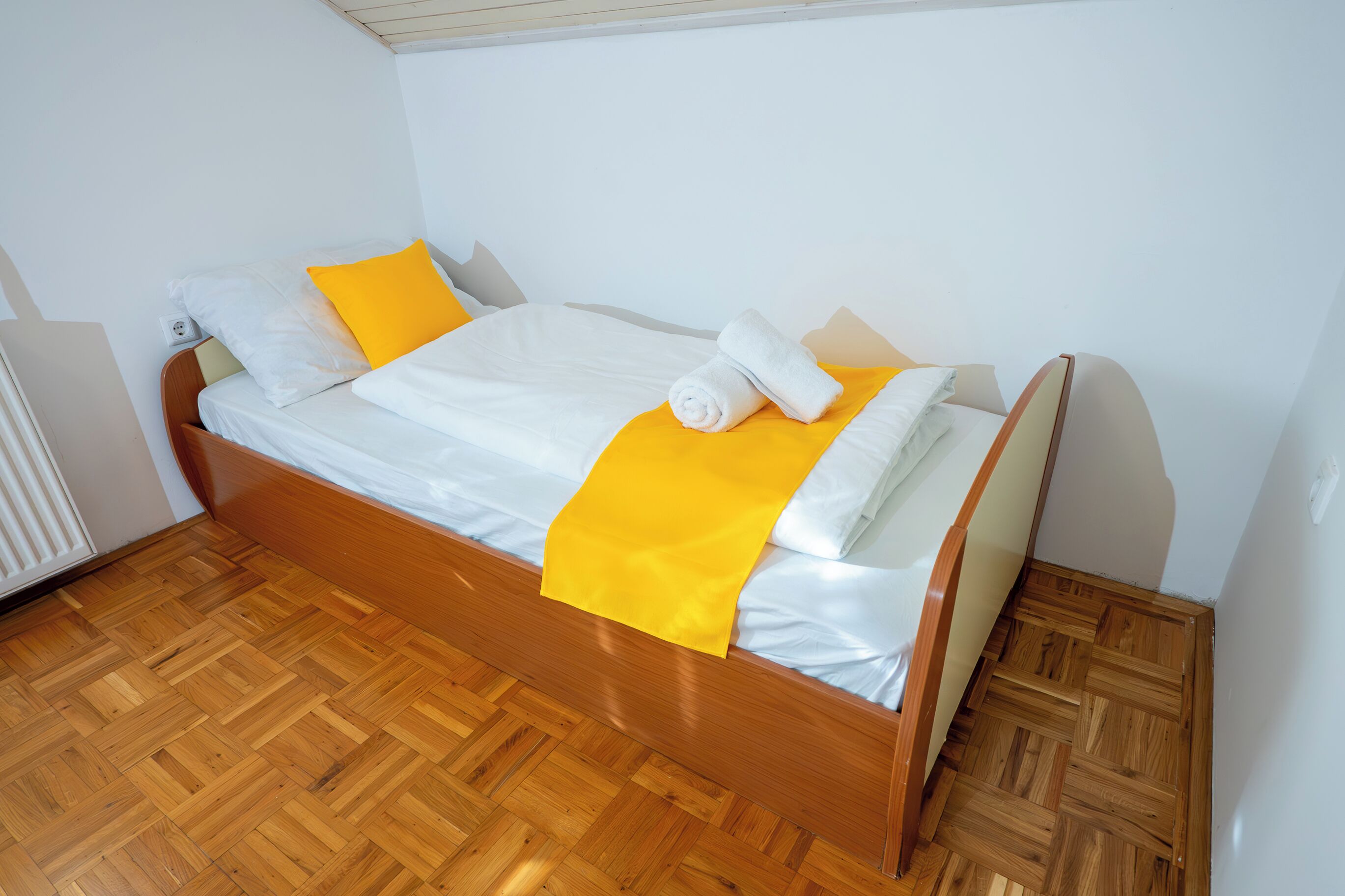 Een eenpersoonsbed met houten frame, wit beddengoed en een gele accentkleur.