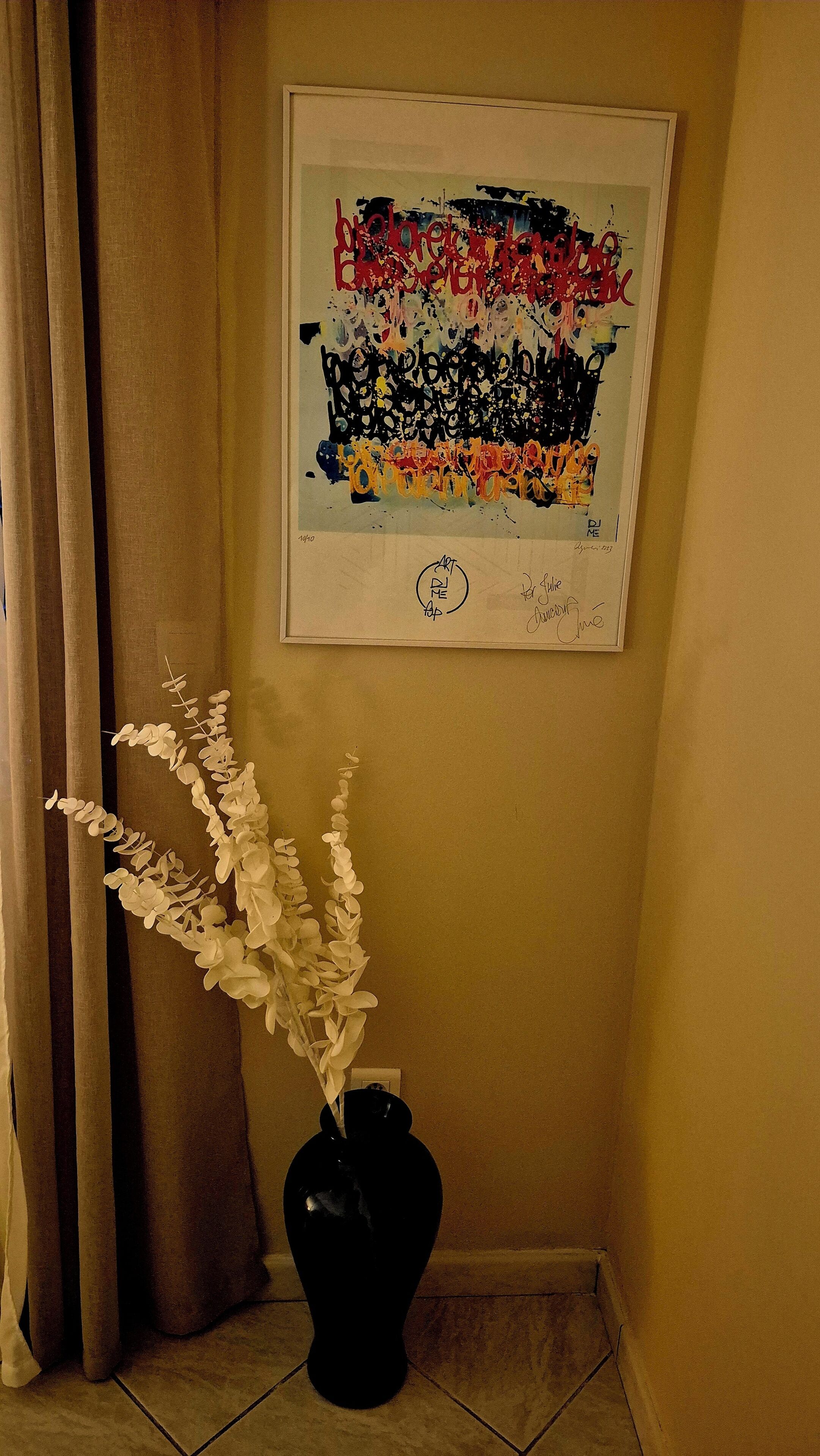 Een lijst met een abstract schilderij aan een muur, een zwarte vaas met witte bloemen op een betegelde vloer, en een gedeeltelijk zichtbaar gordijn.