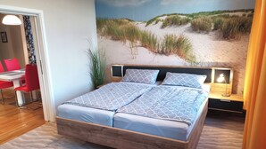 A bedroom with a bed, a nightstand, a lamp, a wall mural of a beach, and a dining area with red chairs and a table.