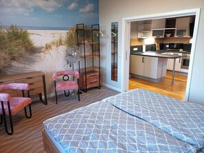Une chambre à coucher avec un lit, une chaise, un bureau, un ventilateur et une vue sur la plage, visible par la fenêtre.